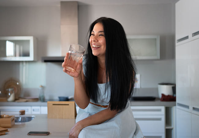 A woman with long, black hair smiling in her kitchen as she takes Brain Boost with a glass of water